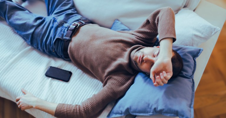 A man lying on a couch with his phone beside him, highlighting Sleep Mistakes After 30 such as late-night screen use that can disrupt sleep quality.